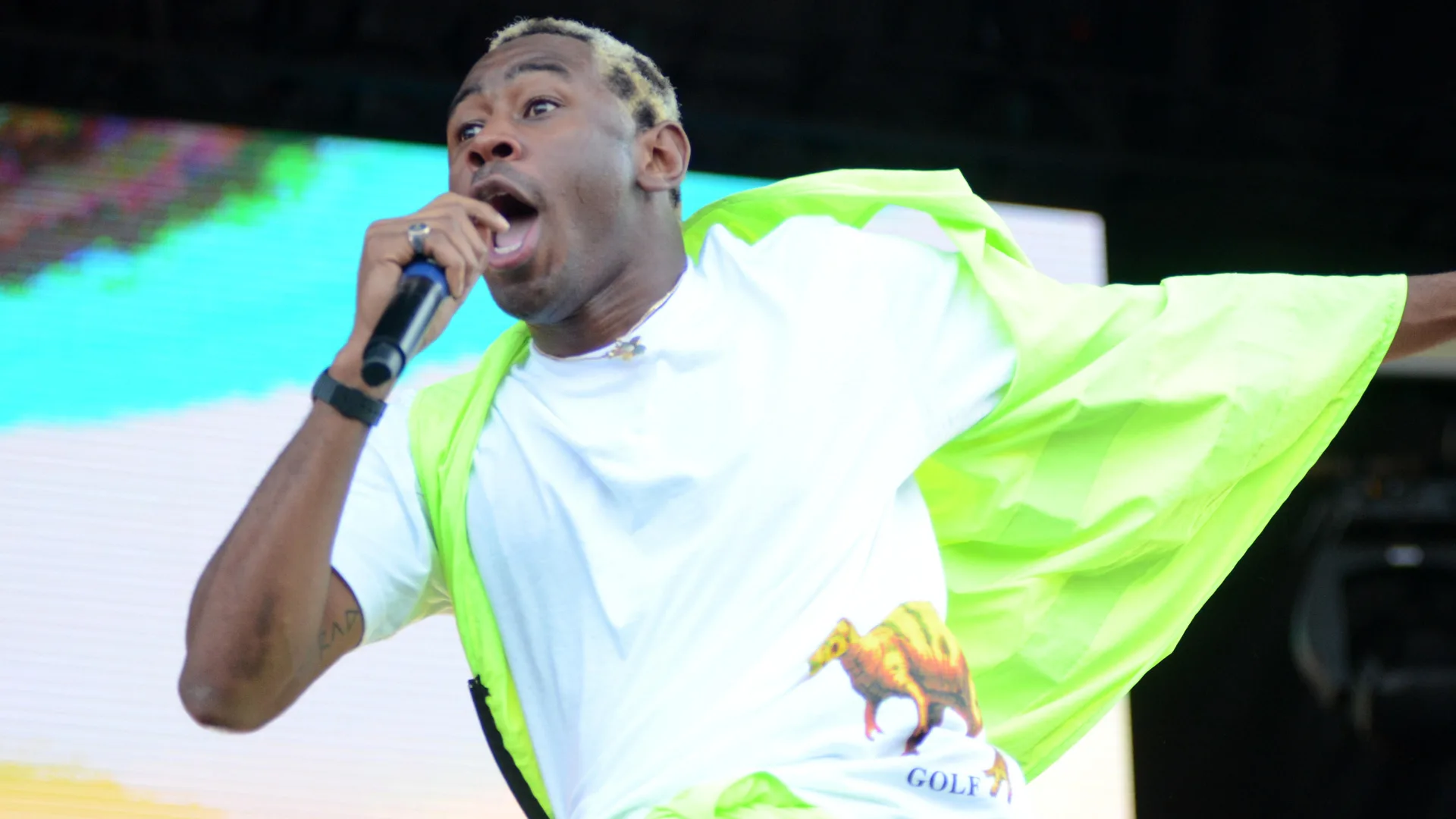 A photo of Tyler the Creator wearing a white top and lime green jacket blowing in the wind singing into a mic on stage with a black white and blue background.