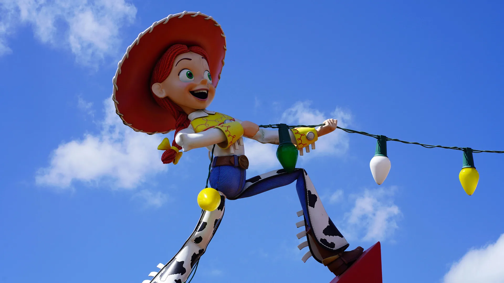 A photo of Jessie the cowgirl from Toy Story pulling on some lights against a blue sky with white fluffy clouds.
