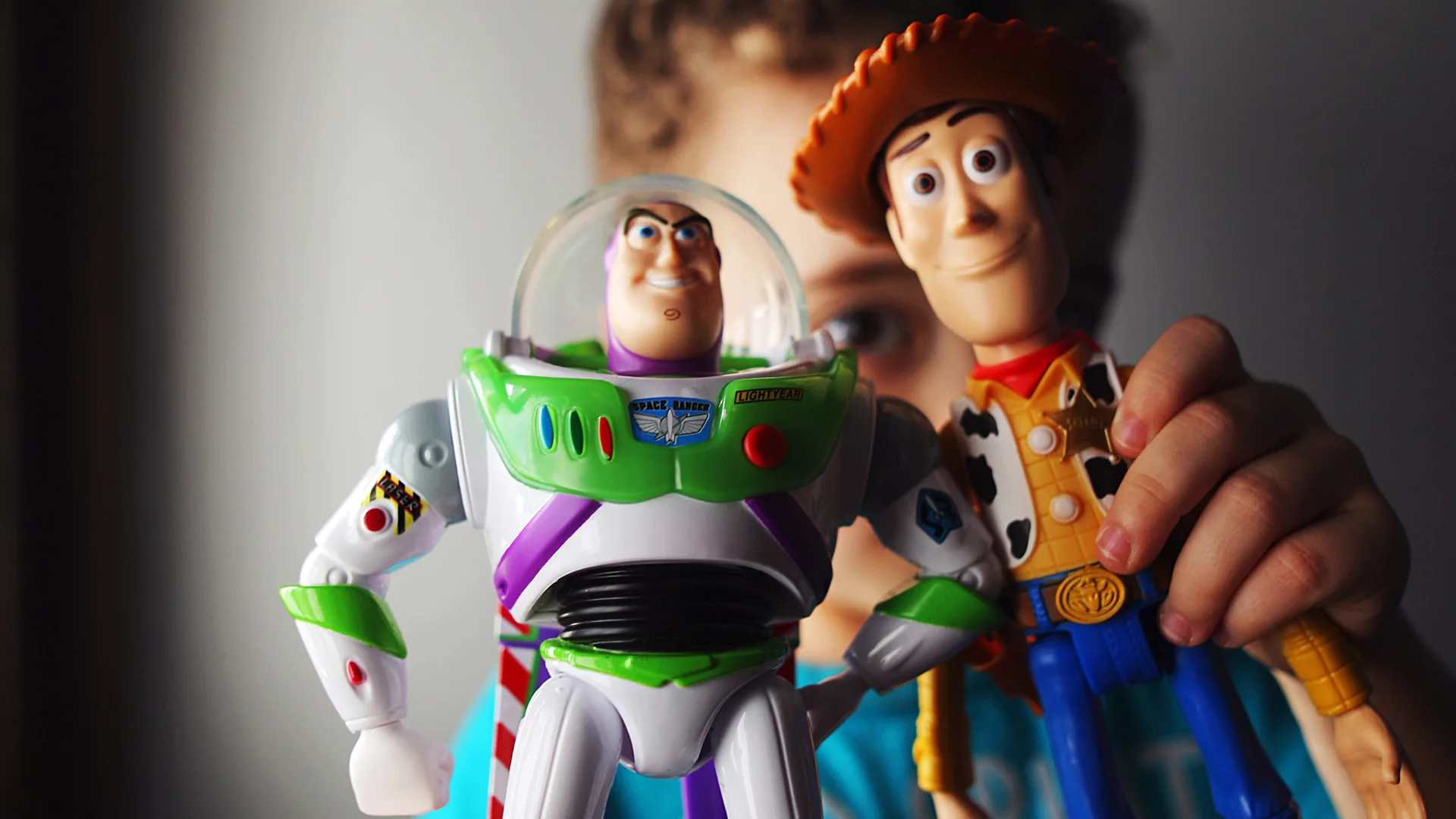 A photo of the Toy Story characters Buzz Lightyear and Woody being held by a child just out of focus against a grey background.