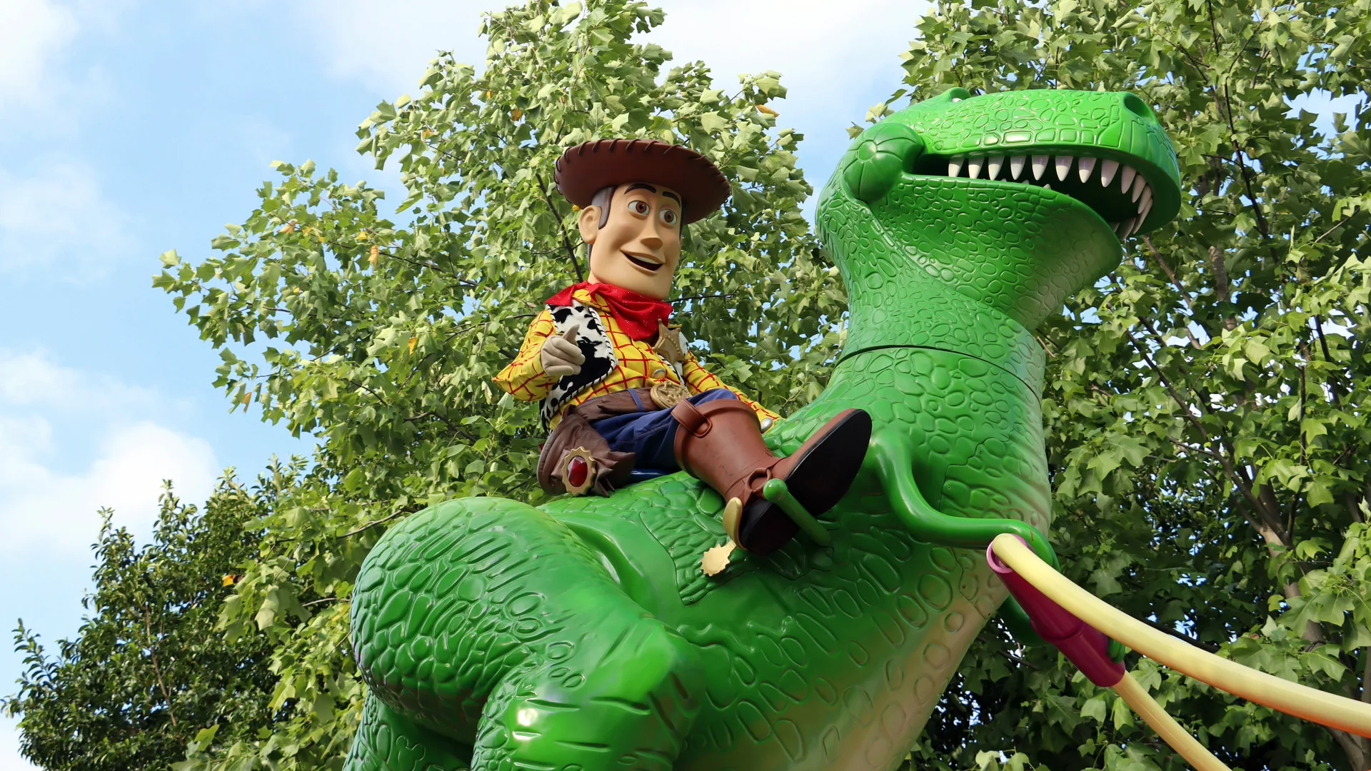 A photo of Woody from Toy Story sat on the back of the dinosaur set against some green trees and blue cloudy sky.