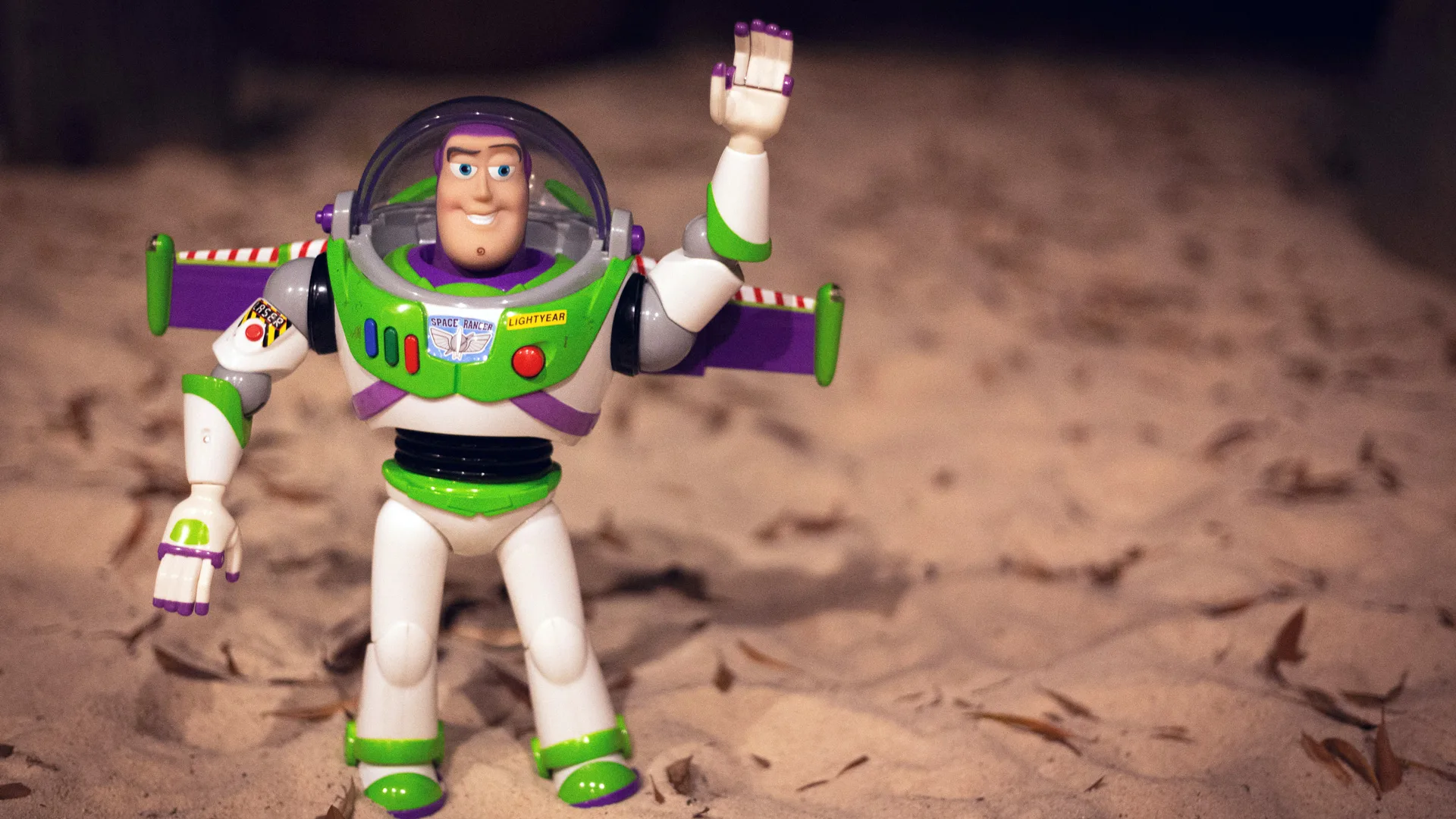 A photo of Buzz Lightyear from Toy Story stood on sand waving one arm in the air.