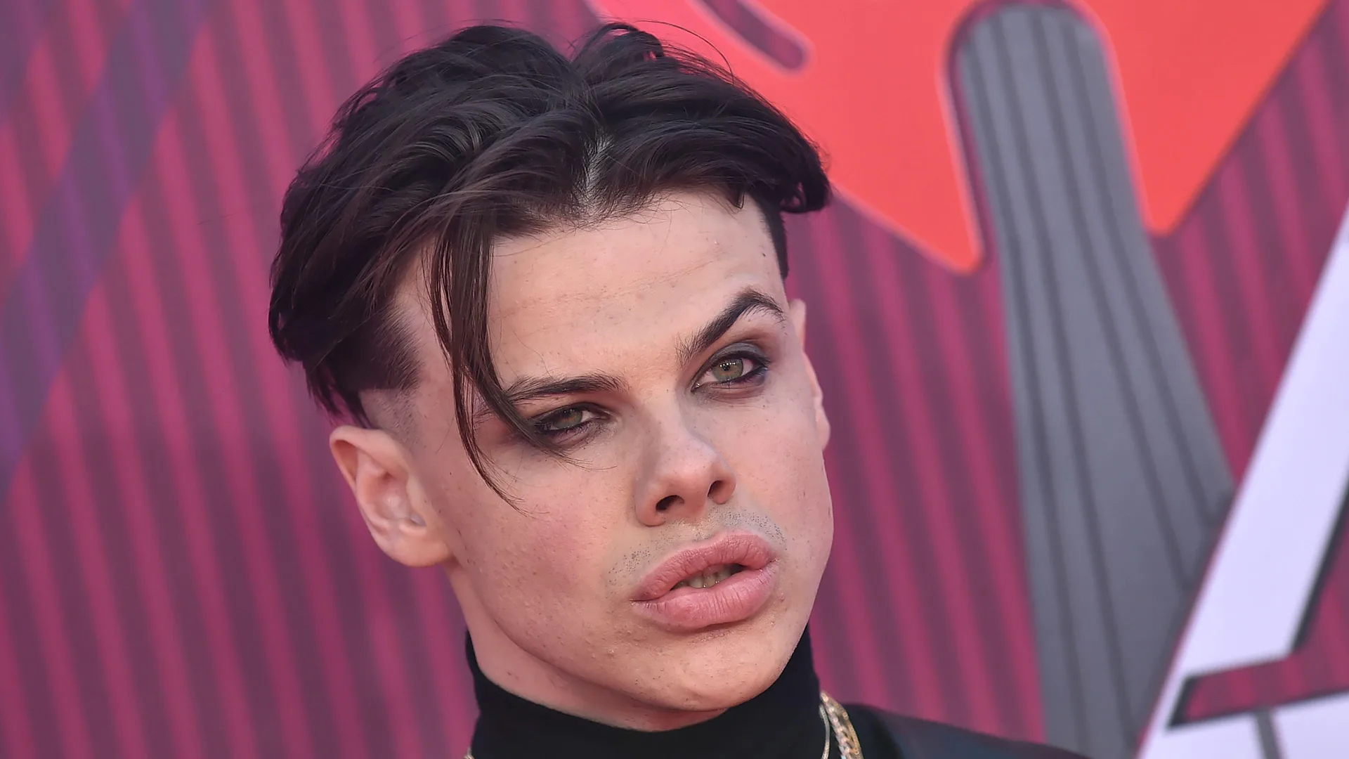 A photo of Yungblud looking into the distance, close up of his face stood against a pink striped background.