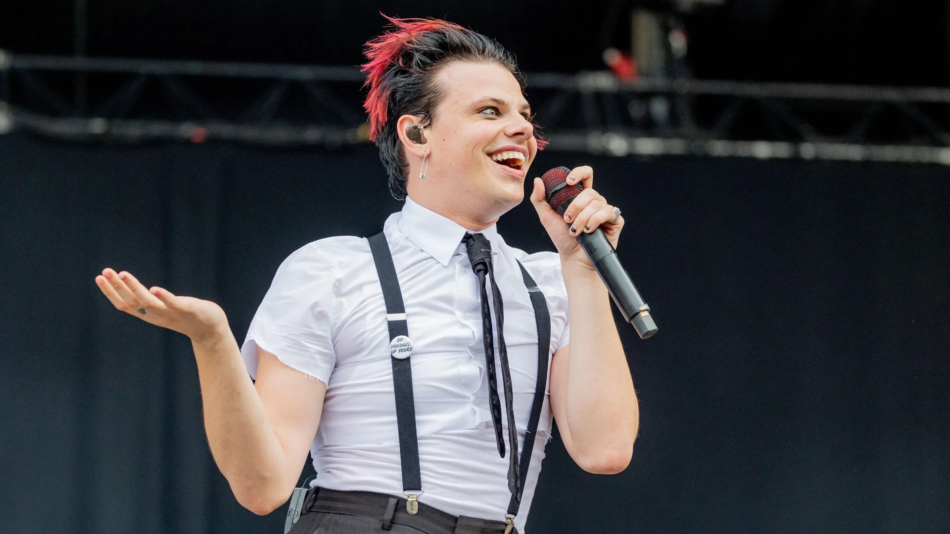 A photo of Yungblud on stage smiling with a mic in his hand and one hand outstretched. He is wearing a white shirt, tie and braces against a dark stage background.