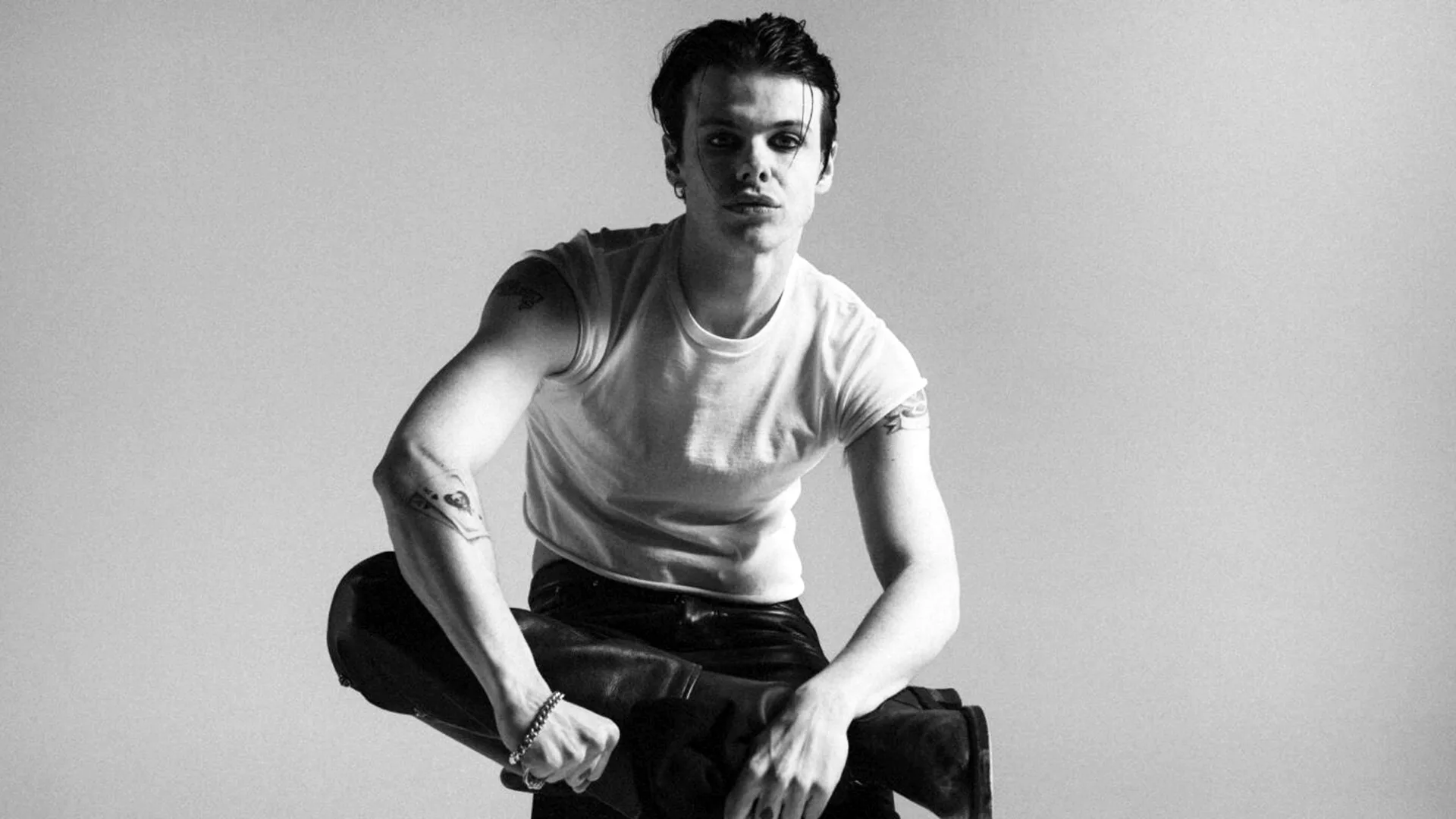A black and white photograph of the singer Yungblud sat with one leg balanced on the other and arms resting over it staring at the camera wearing a white tshirt and black jeans against a grey background.