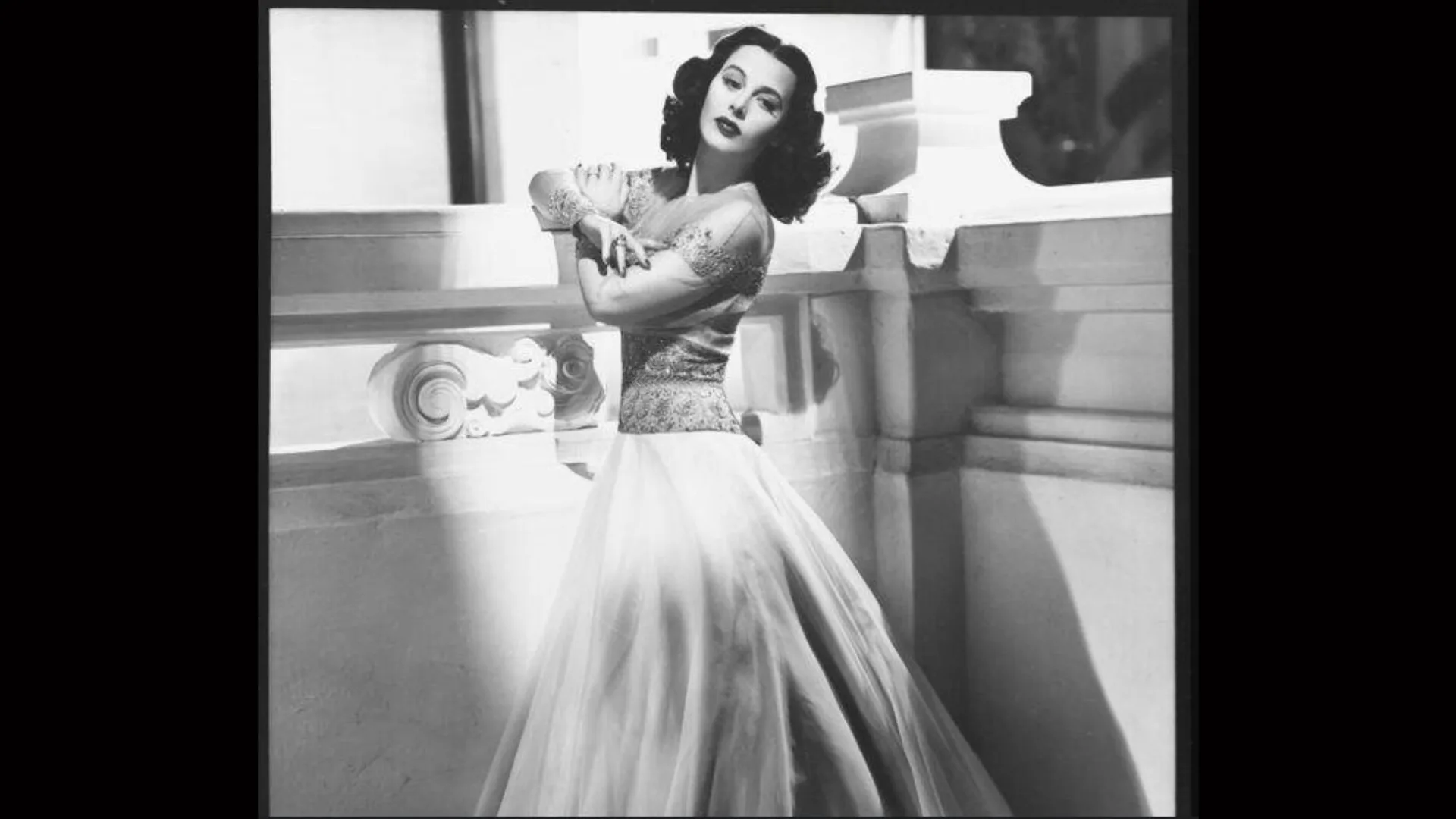 A black and white photograph of Hedy Lamarr stood in long gown with her arms crossed looking over her shoulder - she is stood next to an ornate balcony and pillar with black borders either side of the image.