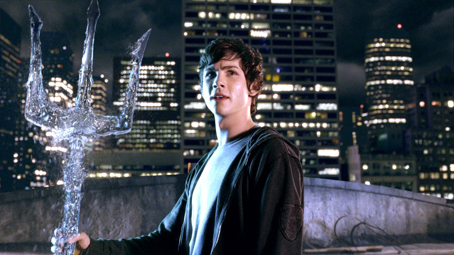 A still from Percy Jackson showing Percy holding a trident - behind him is a cityscape background at night.