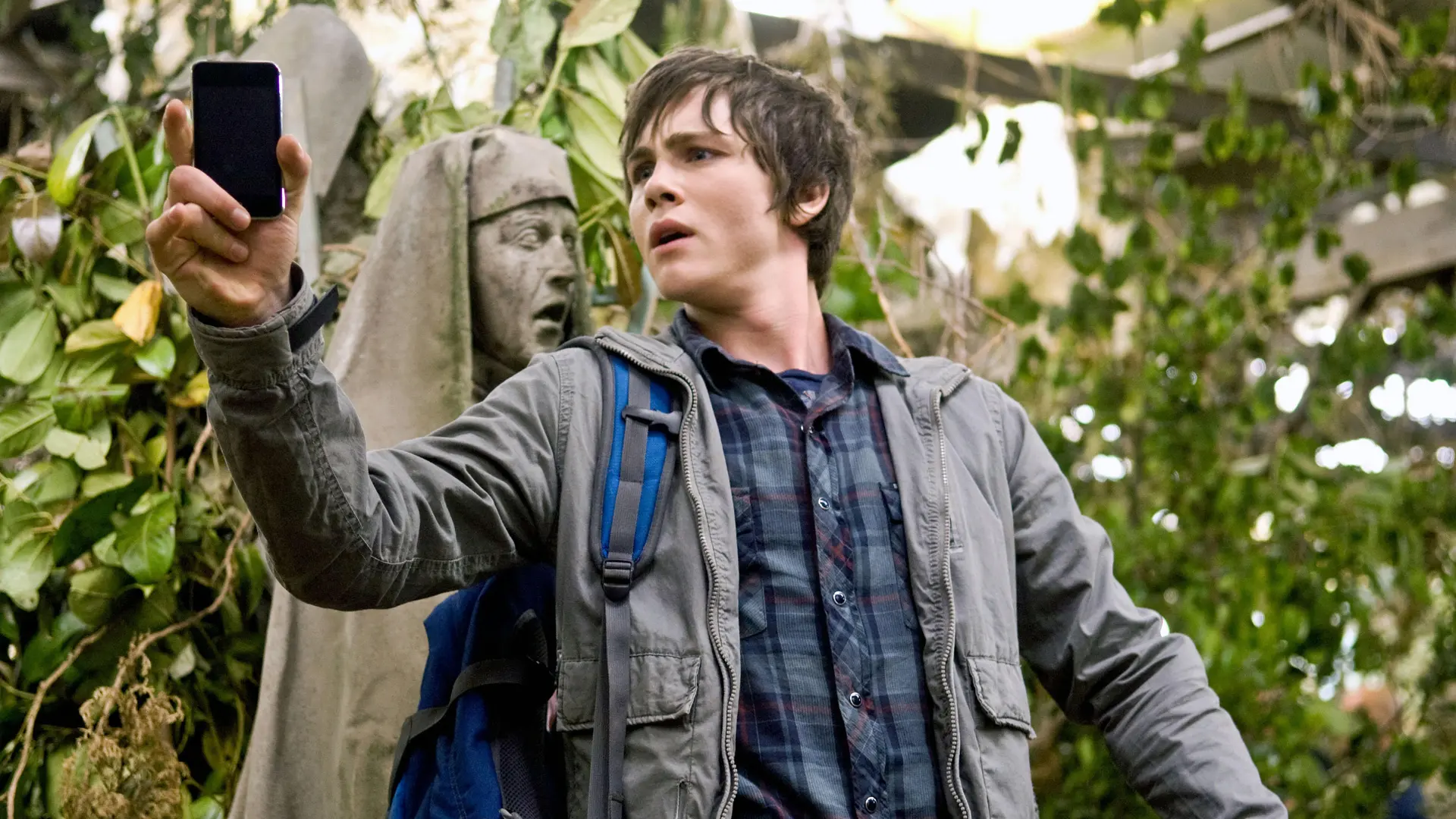 A still from Percy Jackson showing the main character holding a phone looking distressed in the middle of a jungle setting with a statue of a shrouded woman behind him.