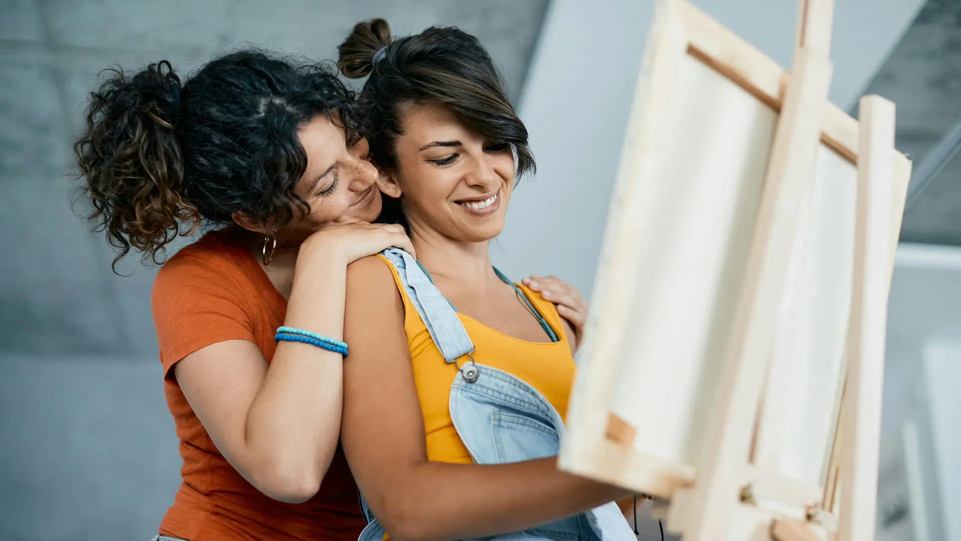 A photo of two women stood at an easel - the two women are closely stood together with one behind putting her hand on her shoulder.