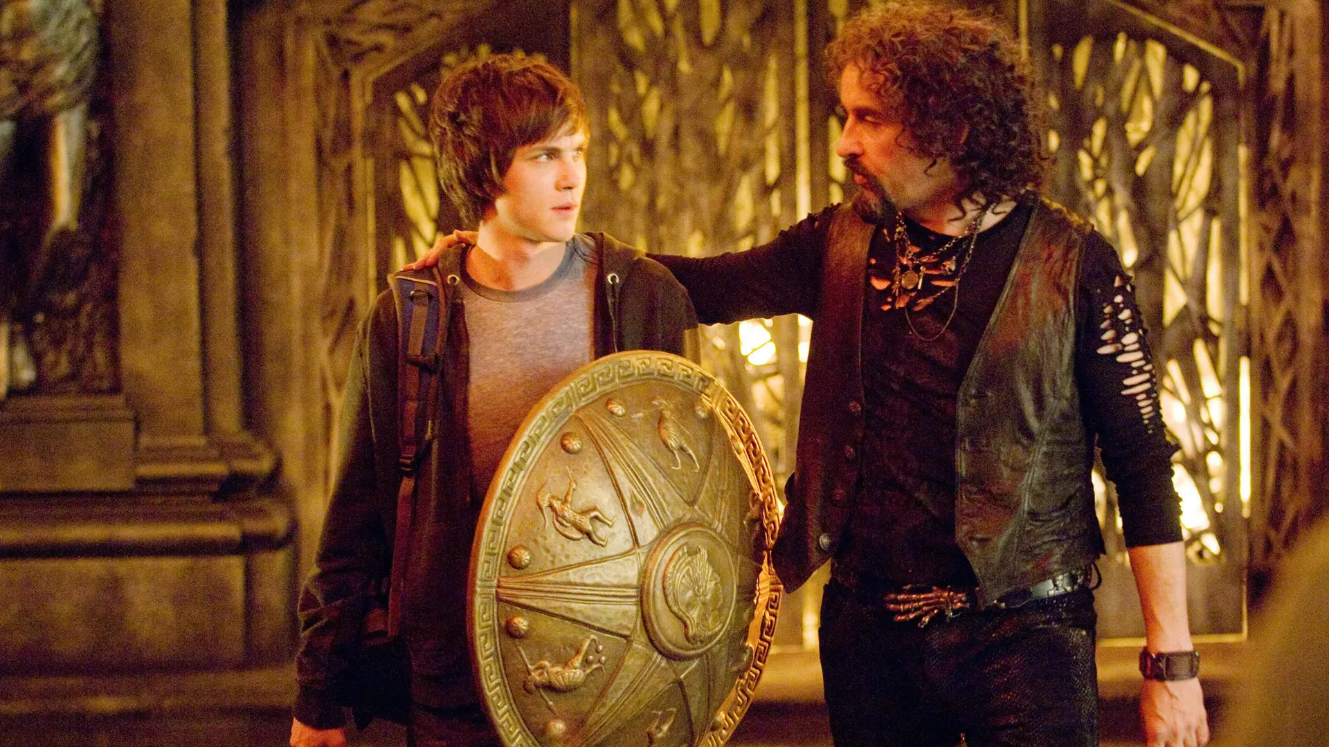 A still from the show Percy Jackson showing Percy holding a golden shield stood next to a man putting his hand on his shoulder in a gold room.