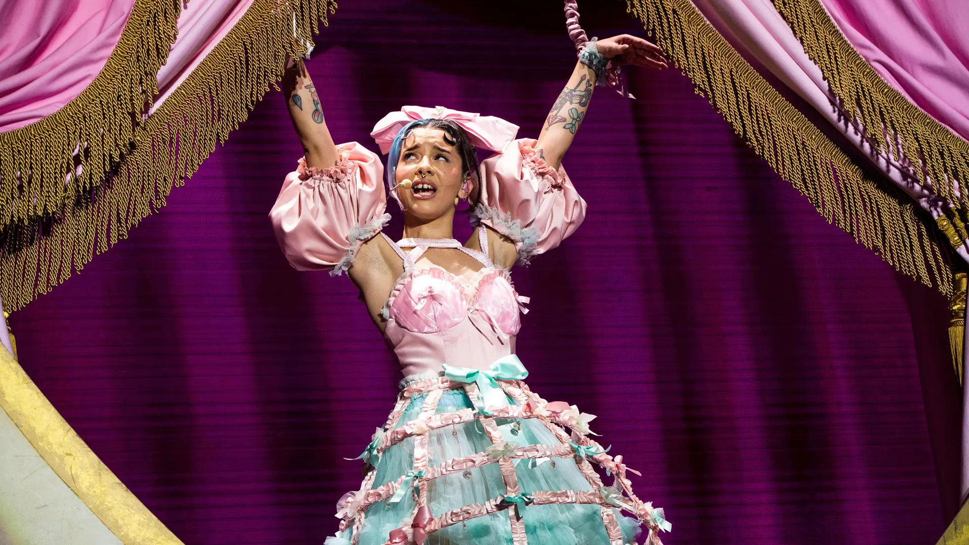 A photo of singer Melanie Martinez with her hands in the air wearing a pink corset and a blue and pink caged petticoat skirt with pink curtains either side of her with gold fringe.