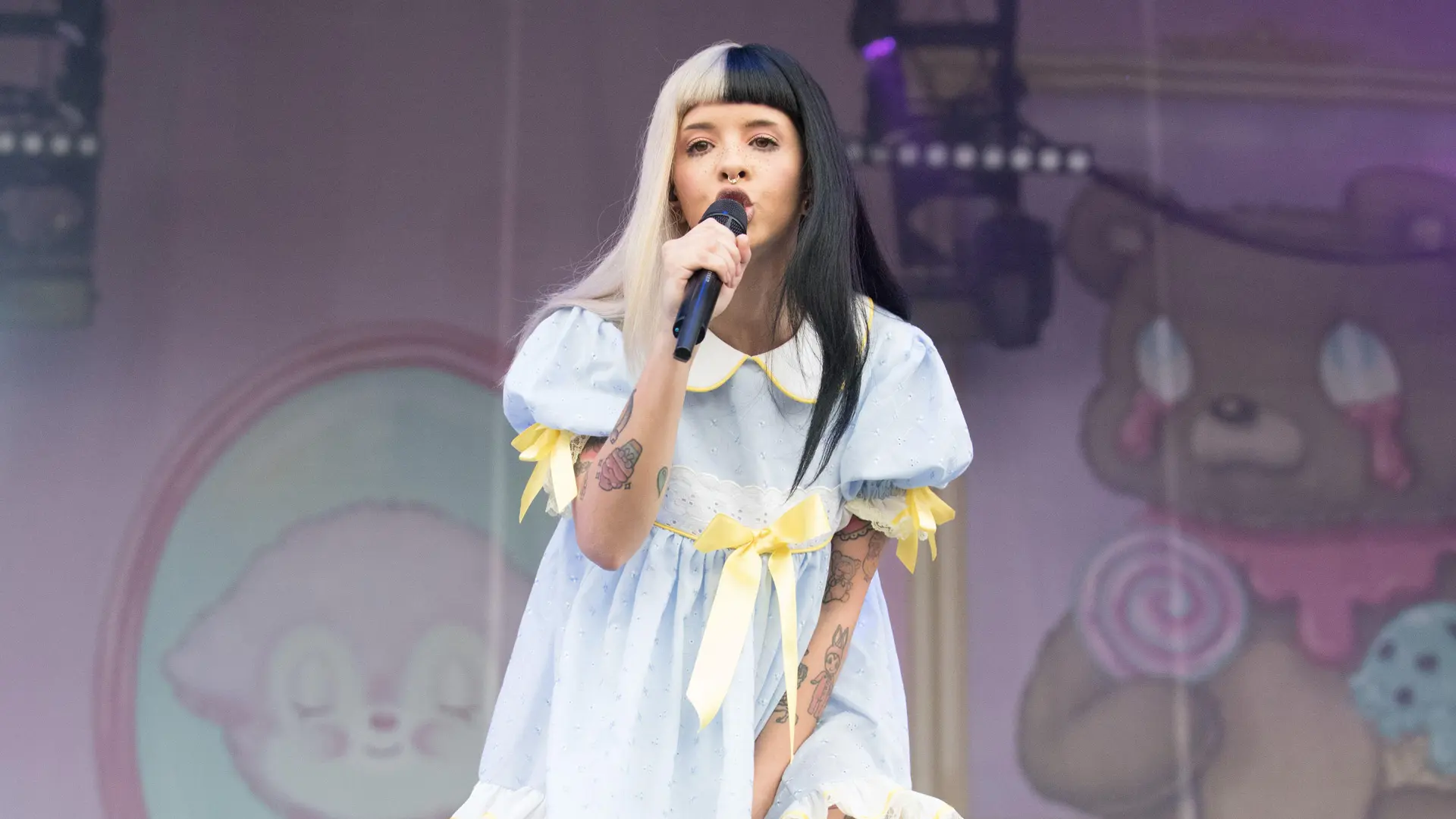 A photo of the singer Melanie Martinez showing her singing into a mic on stage wearing a blue and yellow babydoll dress with black and white hair.