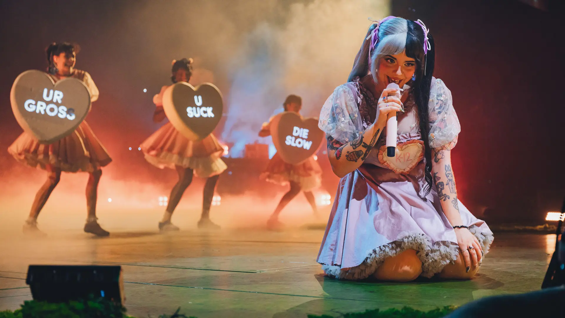 The singer Melanie Martinez on the floor of a stage singing into a mic with three backing dancers behind holding lovehearts with messages and smoke and lights behind them.