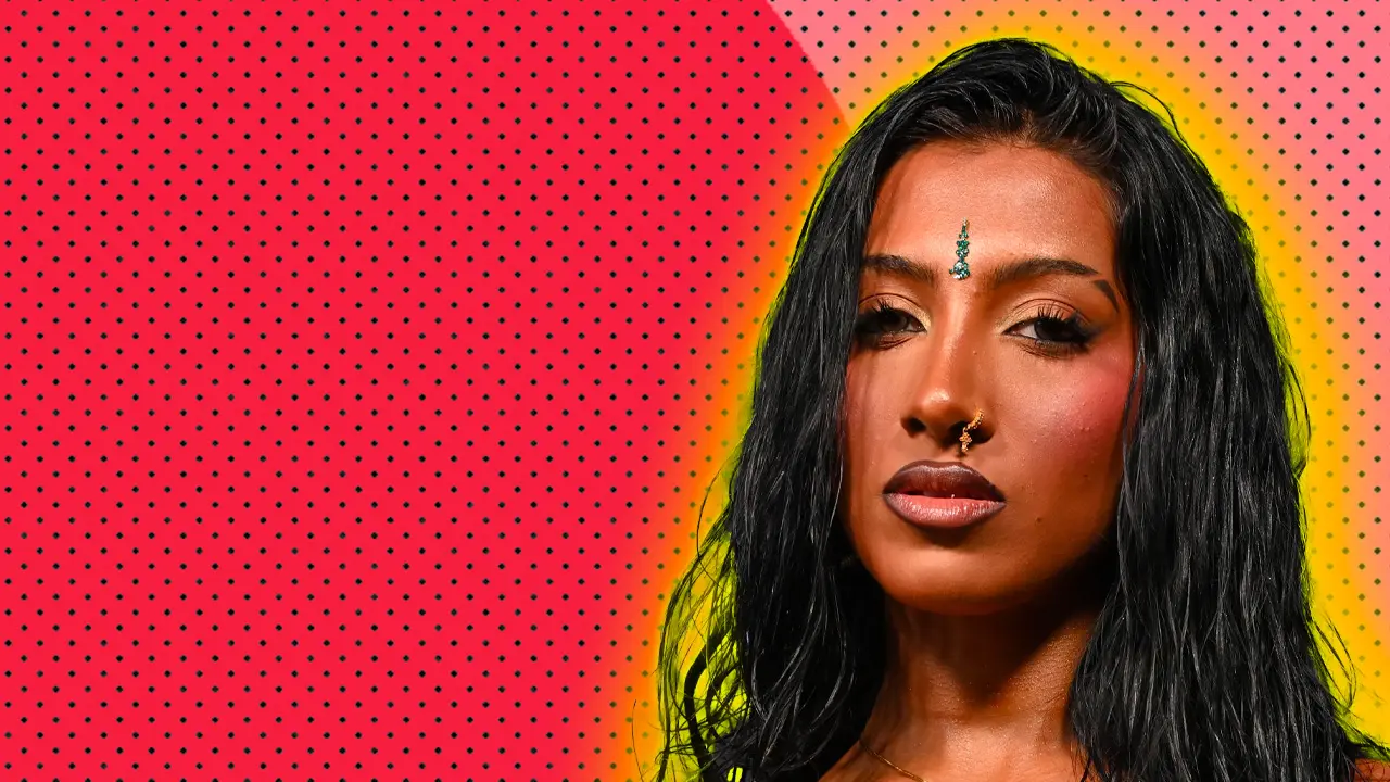A photo of Lara from Katseye looking directly at the viewer with a nose ring and bindhi on her forehead and long hair set against a red and pink dotted background with a yellow halo.
