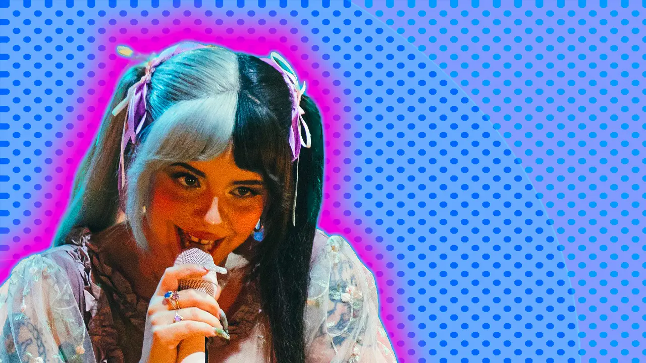 An image of Melanie Martinez smiling into a mic with black and white hair in pigtails with pink ribbons set against a blue dotted background with a pink halo
