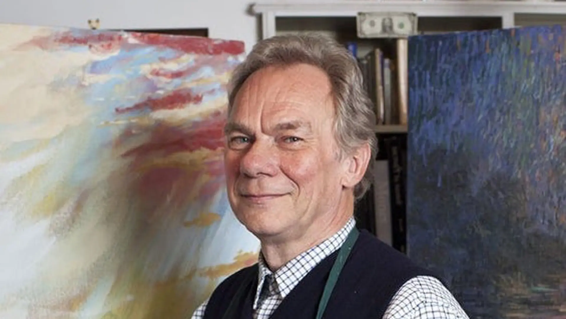 A photograph of John Myatt smiling directly at the camera with a painting behind him