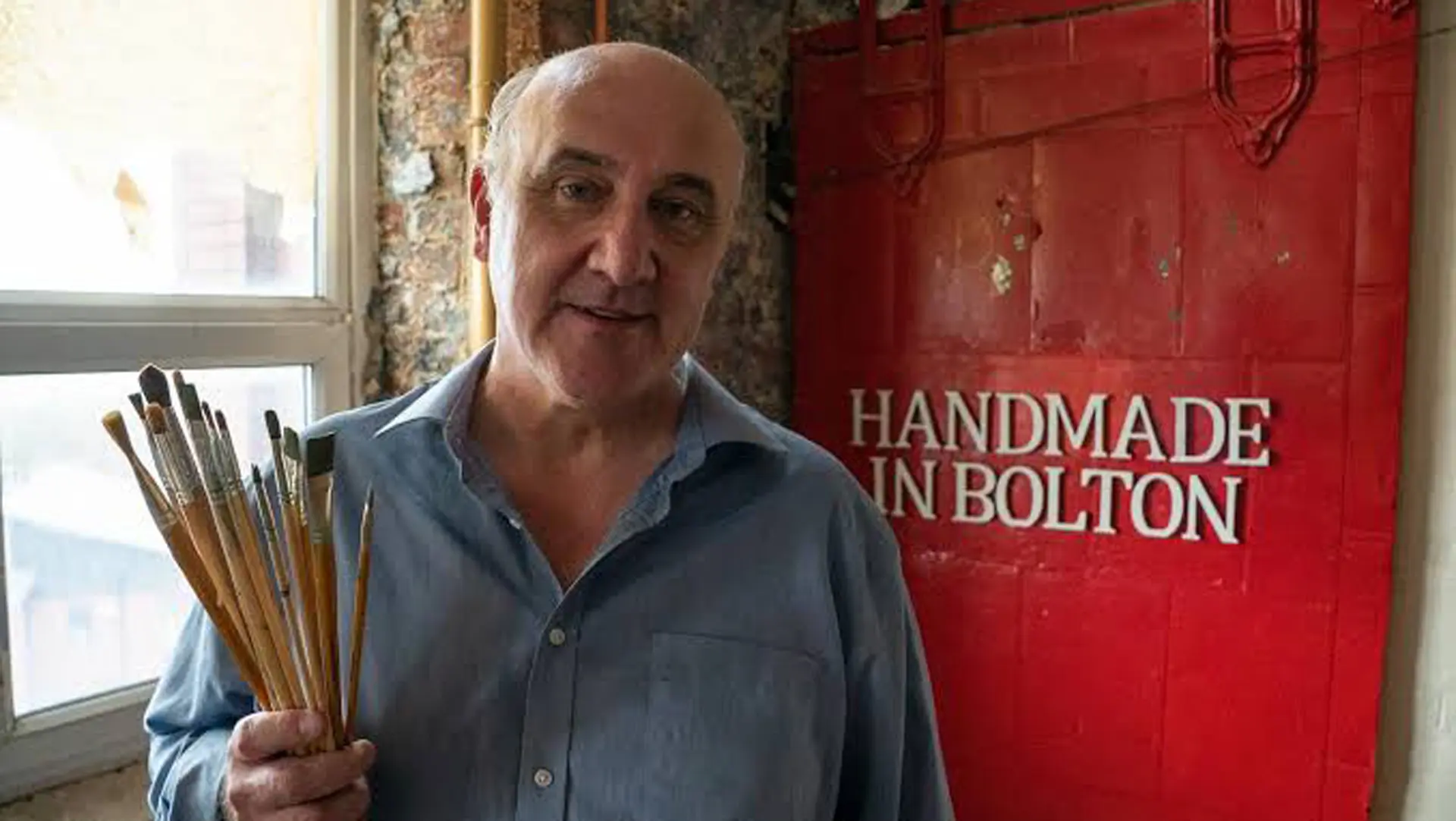 A photo of Shaun Greenhalgh, part of the Bolton Forgerers - he is stood holding some paintbrushes wearing a blue shirt next to a window and a red sign that says HANDMADE IN BOLTON