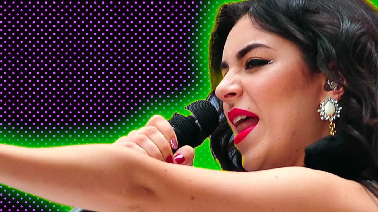 A photo of Charlie XCX singing on stage pointing her arm out waving her finger. She is wearing a zebra print top against a purple dotted background with a green halo.
