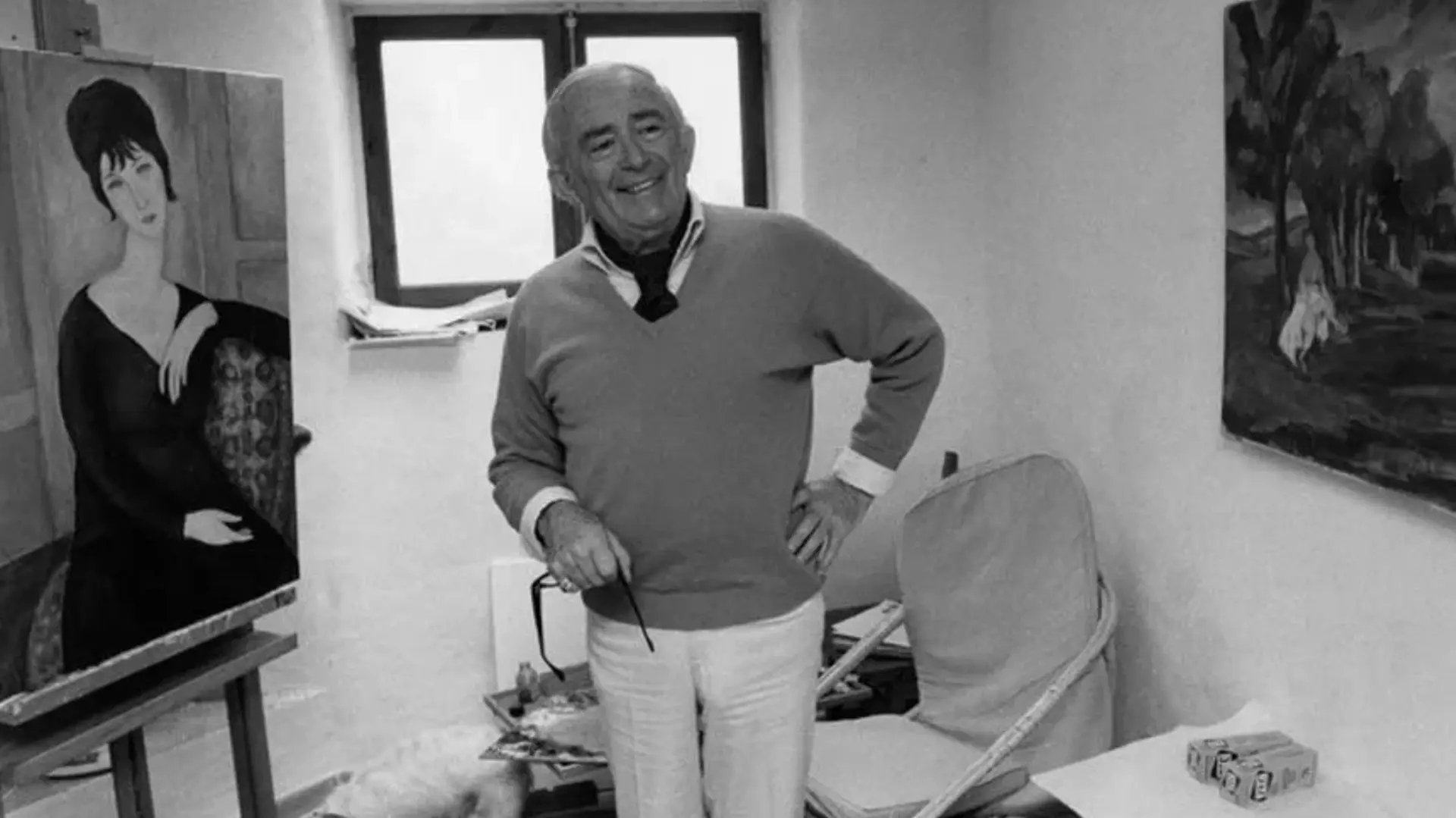 A black and white photo of Elmyr de Hory showing him stood in a room with one of his forged artworks to his right and a chair and little white dog behind him with a window behind his head. He is in a relaxed pose smiling with one hand on his hip.