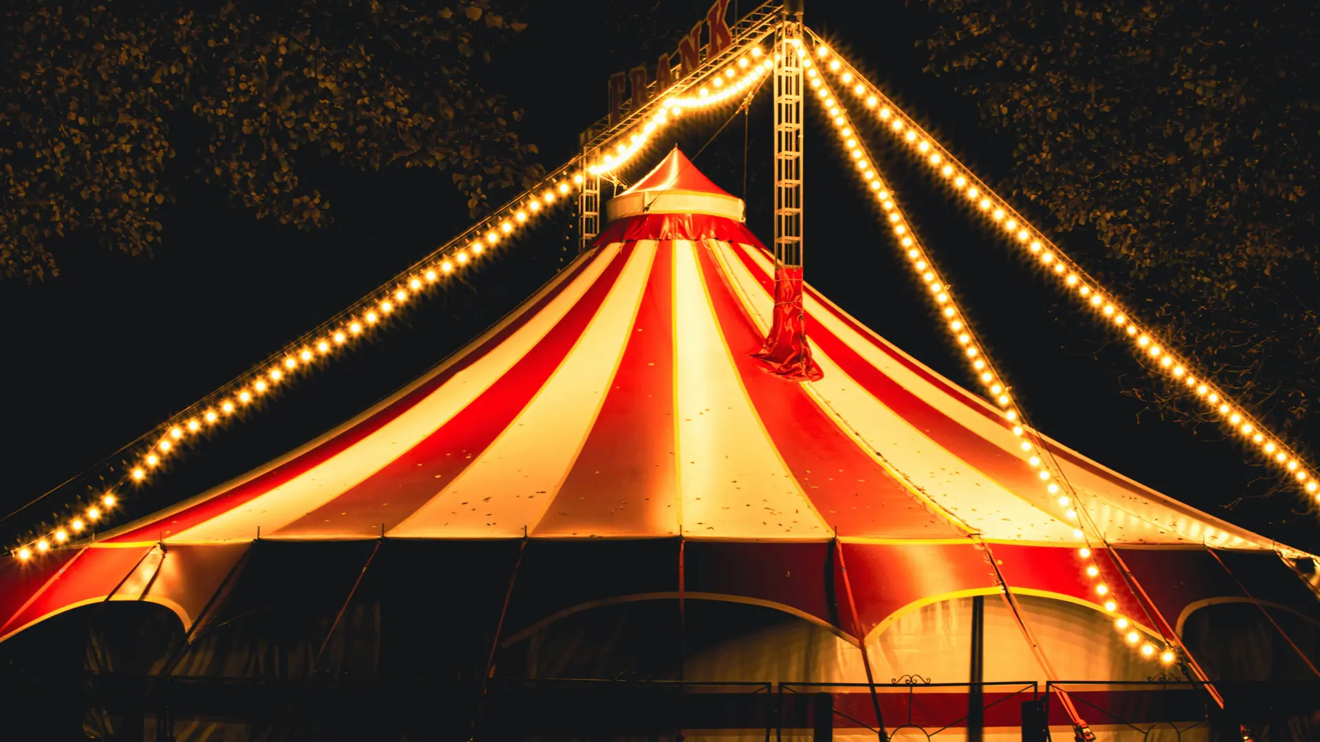 A photo of a circus tent at night all lit up showing the red and white top with festoon lighting coming down the outside.