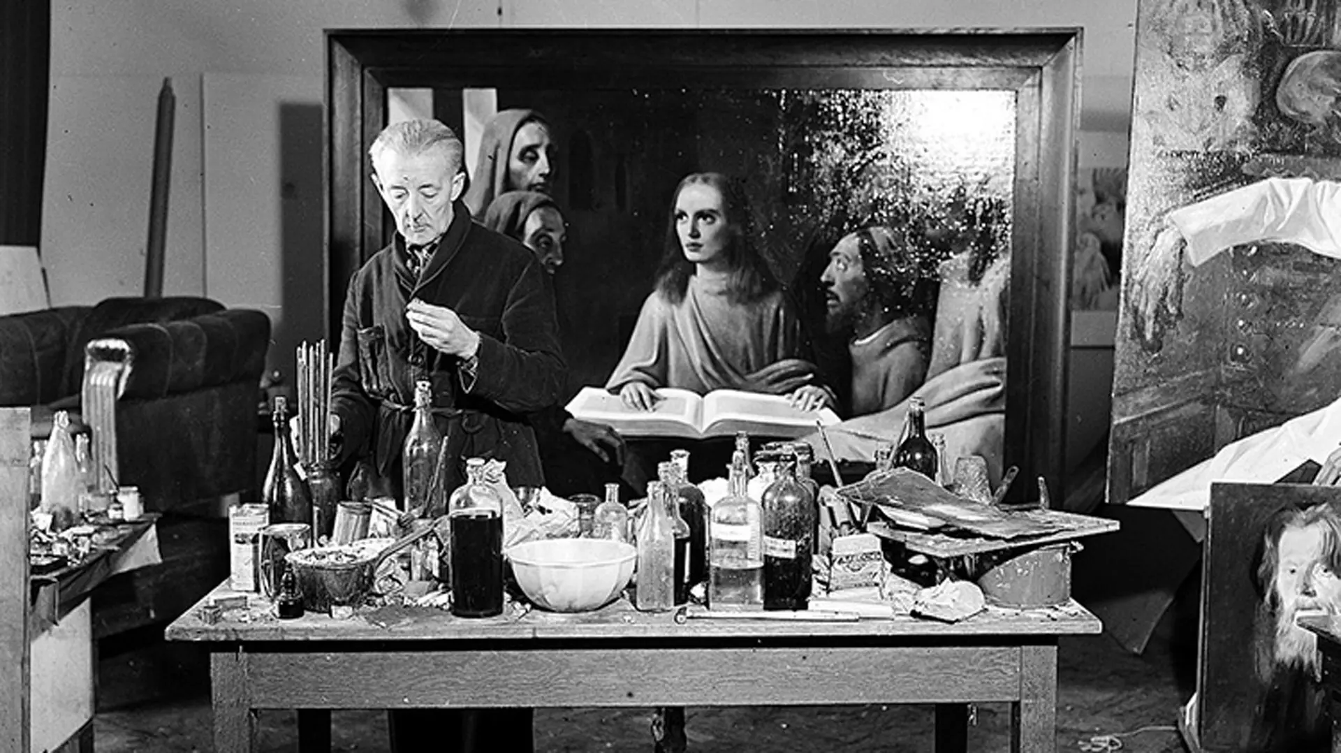 A black and white photo of Van Meergeren in his studio stood over a table of bottles and paints with one of his forgeries behind him depicting a religious scene.