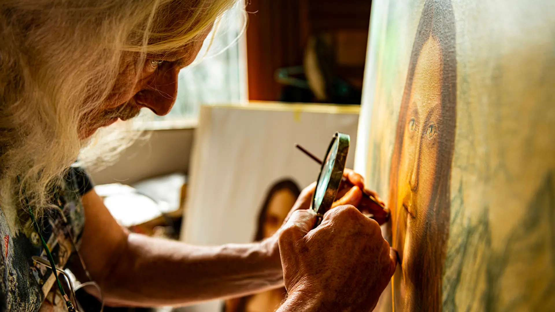 A photograph of Wolfgang Beltracchi showing him holding a magnifying glass and paintbrush to one of his paintings in a soft lit room with paintings behind him and a window with curtain.