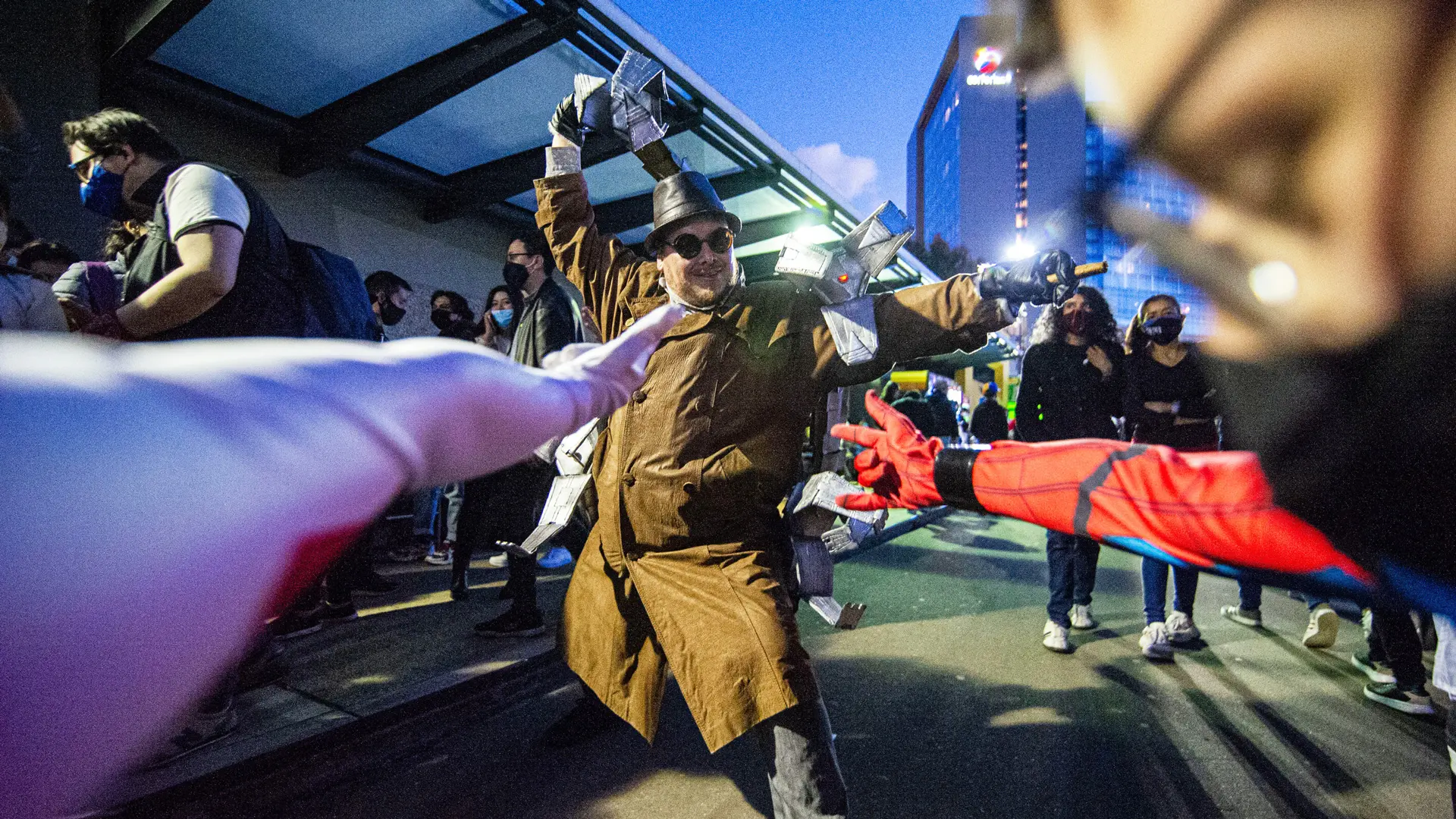 An image of a man dressed as Dr Octopus from Marvel's Spiderman with arms shooting in front of him as he raises his robotic arm in the air against a building and blue sky.