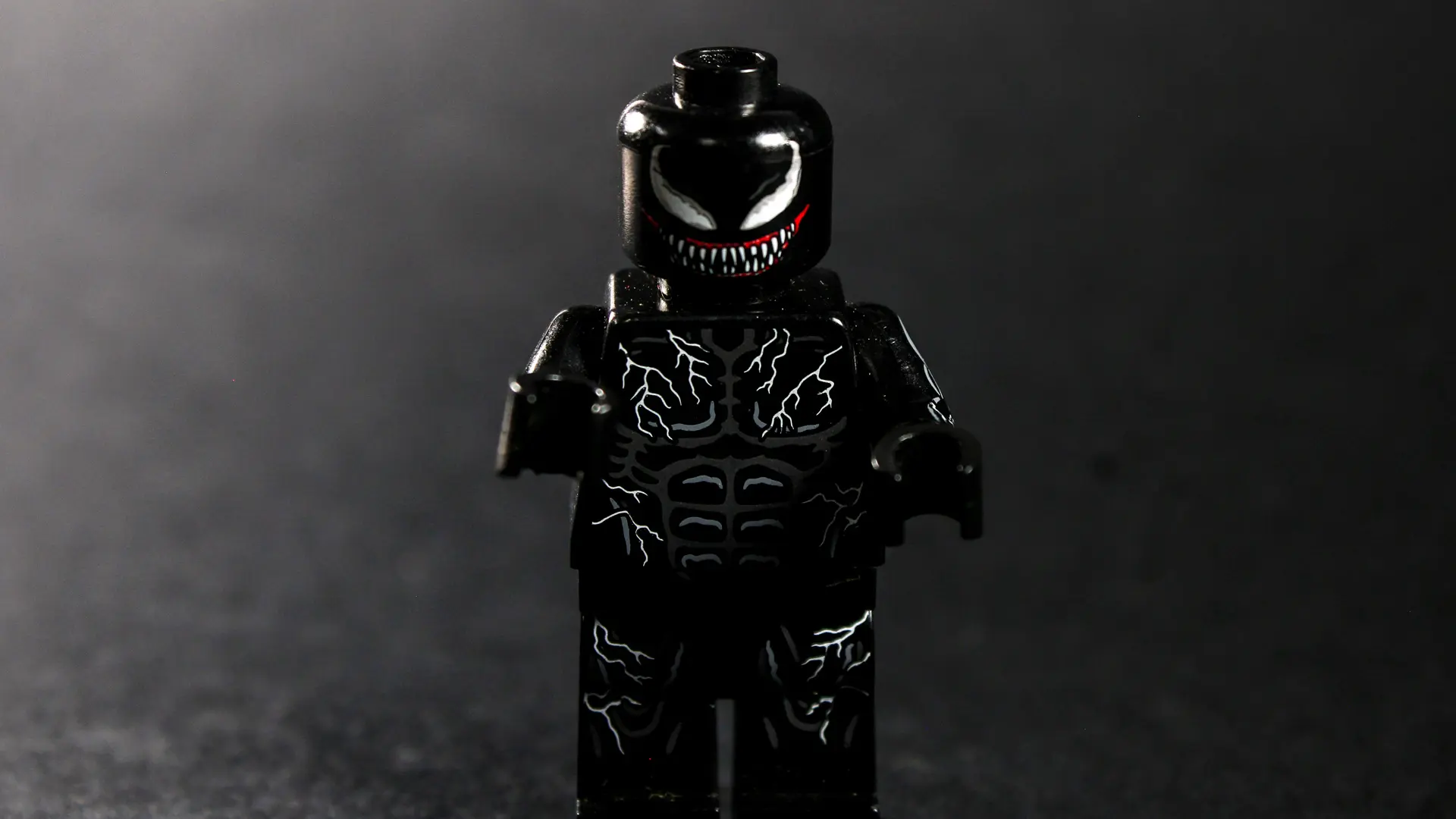 An image of Spiderman Venom with his evil face smiling at the viewer with fangs set against a dark background.