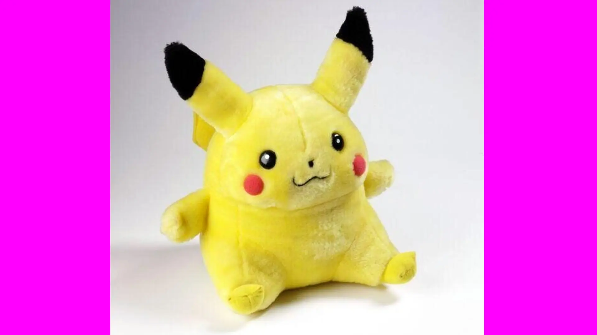 A photo of a stuffed toy of Pikachu, the Pokemon. He is sat down with his little arms spread out against a grey background with pink borders either side.