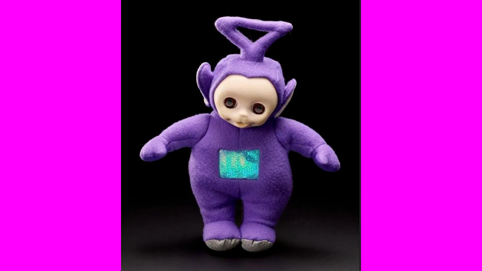 A photo of a purple stuffed toy with a triangular antennae of the Teletubby, Tinky Winky showing him stood up with his arms out against a black background with pink borders either side.