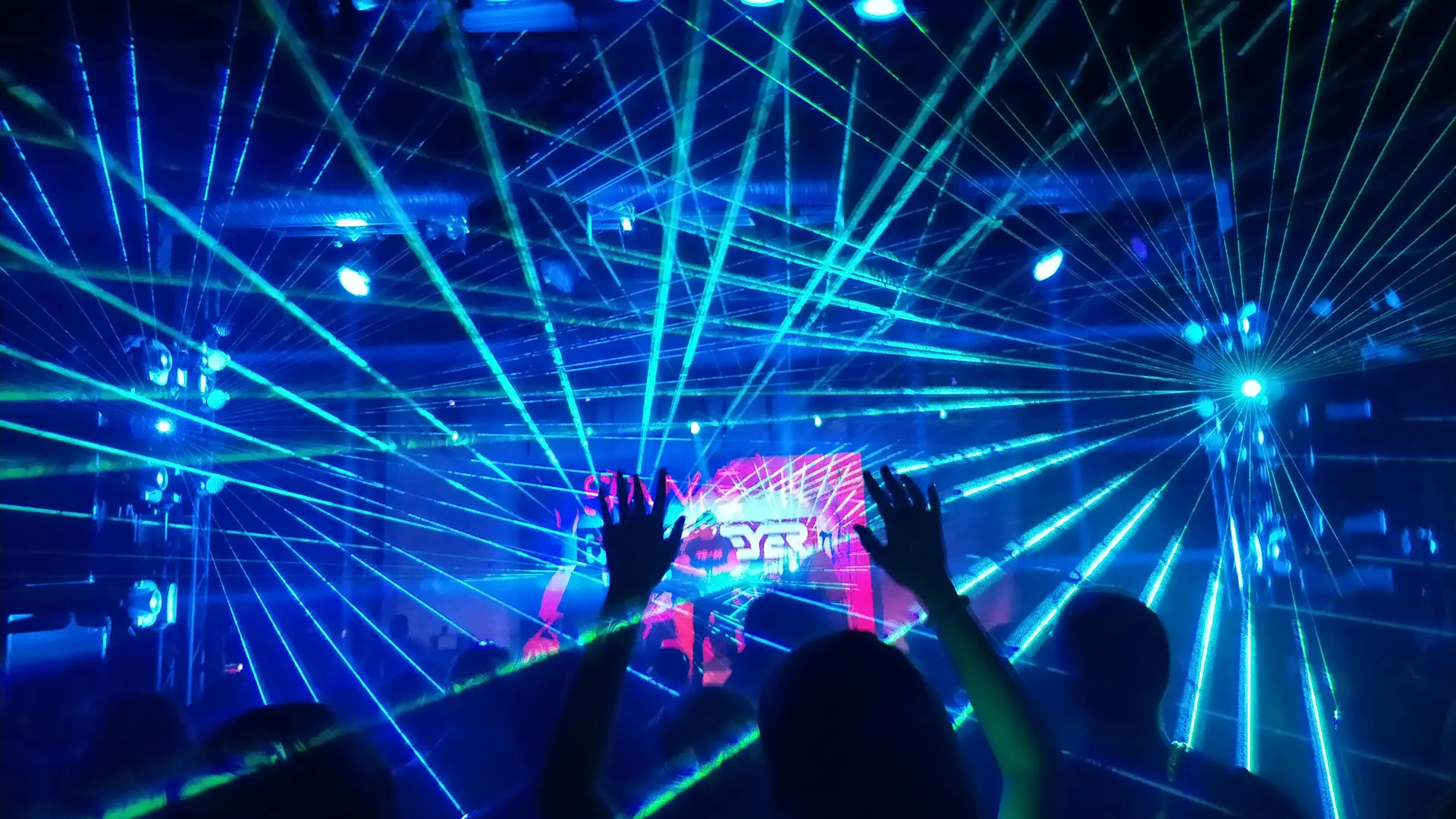 An image of the inside of a club with blue and green laser lights bursting out everywhere and a pink screen in the background with silhouetted hands waving in front.