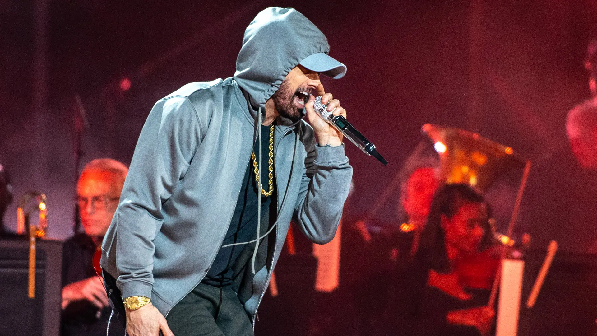A photo of Eminem performing on stage singing into the mic wearing a grey hoodie and cap with red lights in the background.