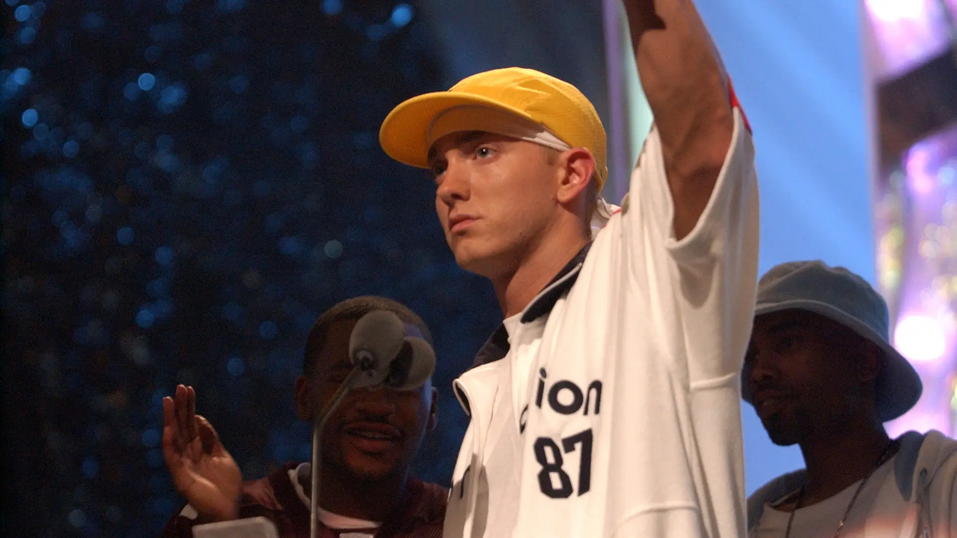 A photograph of Eminem holding his arm up wearing a white shirt and yellow baseball cap with two men behind him.
