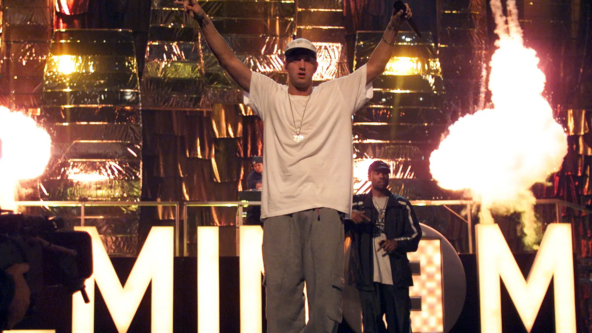 A photo of Eminem on stage with both arms up holding a mic in a white tshirt and grey trousers with the words EMINEM lit up behind him and a person stood watching.