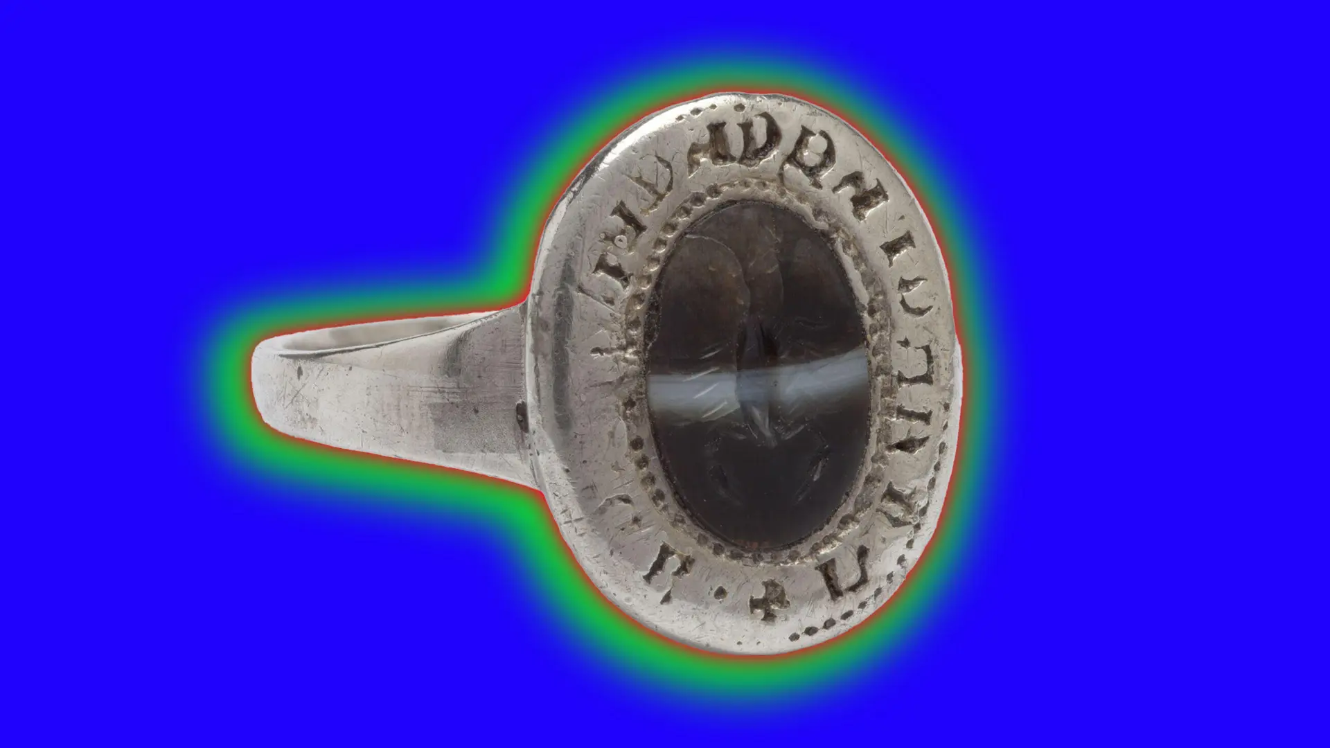 A photo of a silver ring with writing around the edges and a dark engraved centre set against a blue purple background with a red and green halo.