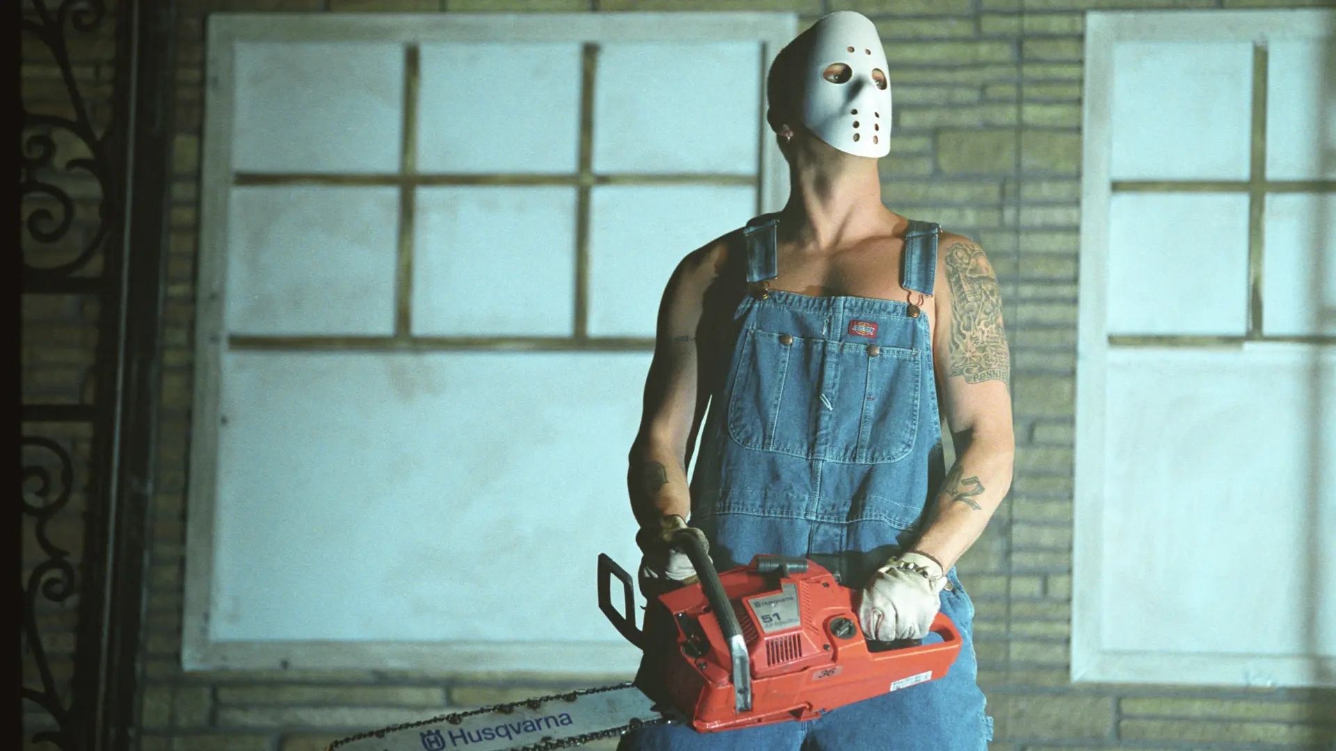 A photo of Eminem in costume with a white hockey mask, blue dungarees and white gloves holding a chainsaw in a room with painted windows on the wall in the background.
