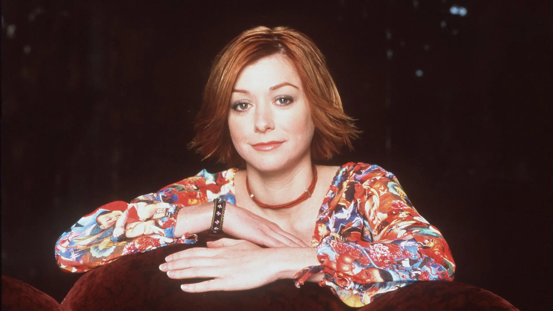 Willow from Buffy the Vampire Slayer sat with her hands over a chair wearing a paisley floral top and smiling at the camera against a black background.