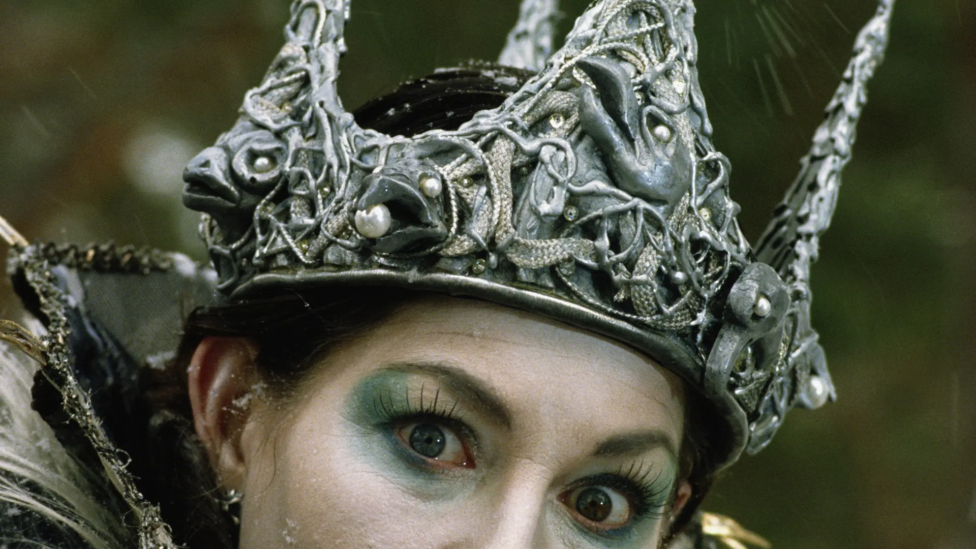 A close up shot of a woman wearing an ornate intricate silver crown with blue and silver eyeshadow on her eyes and dark hair.