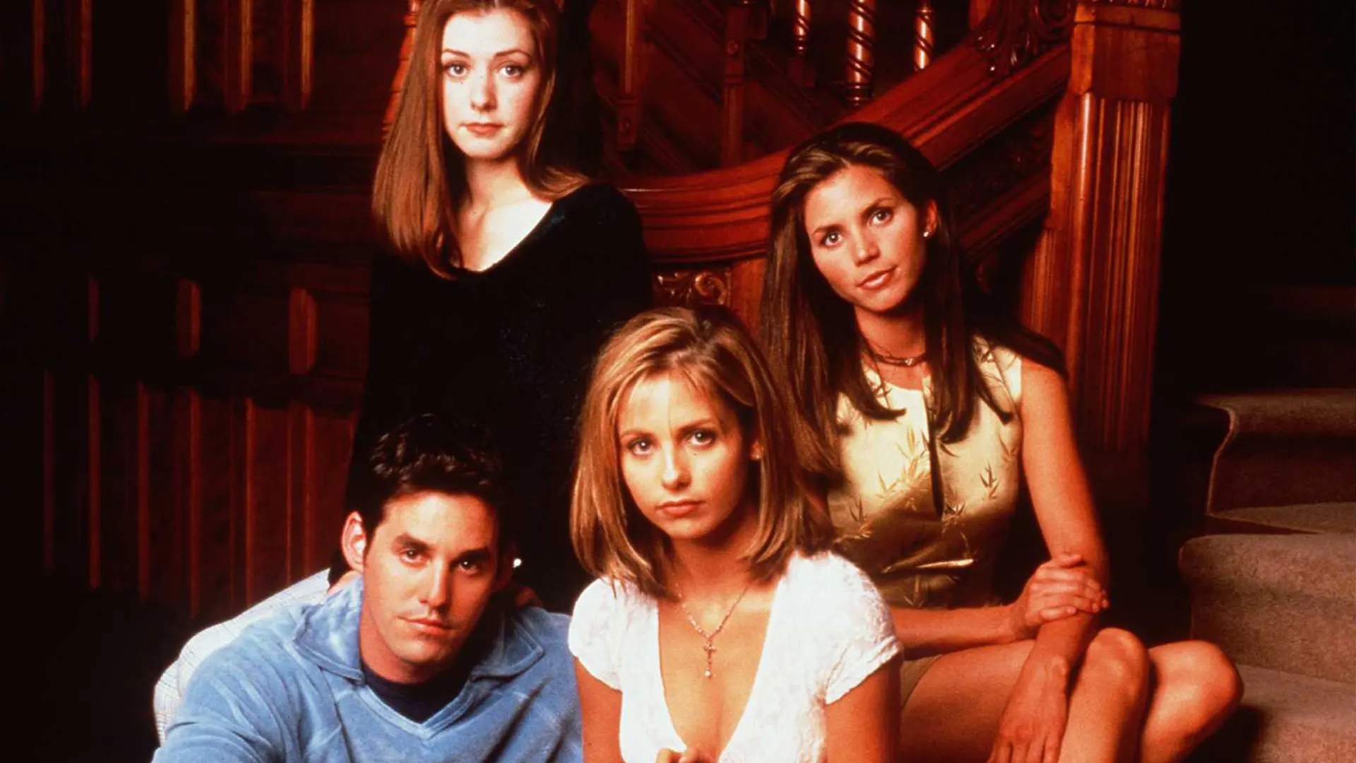 A photo of the cast of Buffy the Vampire Slayer sat on a staircase looking at the camera.