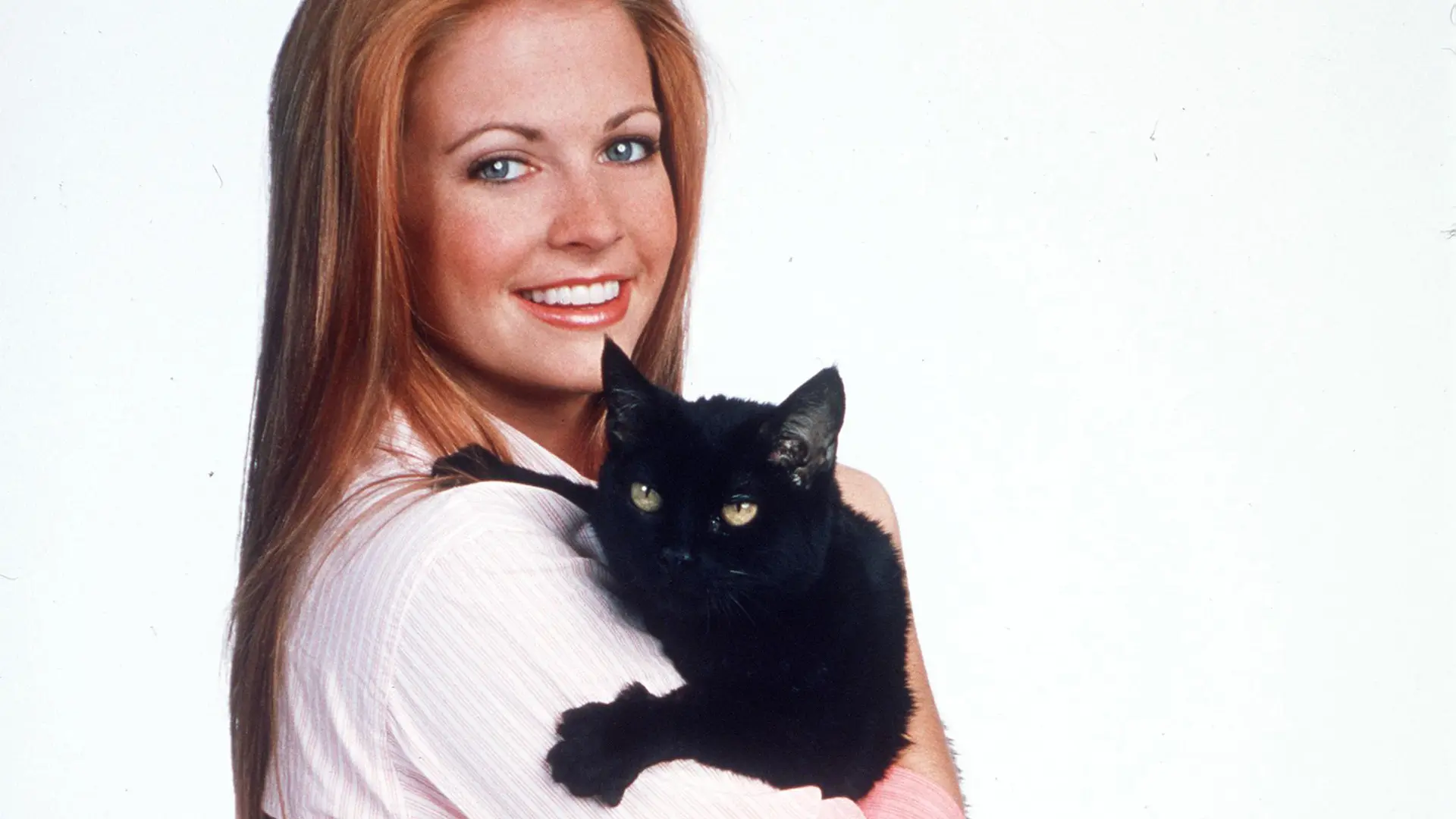 A photo of Melissa Joanhart as Sabrina the Teenage Witch holding her black cat Salem whilst wearing a white top set against a white background.