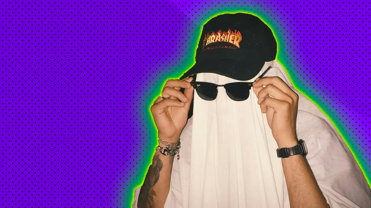 A photo of a man in a white ghost sheet wearing a black cap and sunglasses being held up by his tattooed arms set against a purple dotted background with a green and yellow halo.