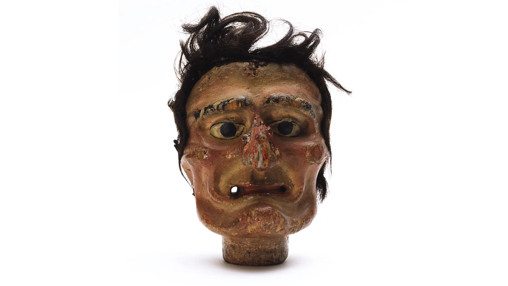 A photo of a hideous looking puppet head with a missing nose, messy black hair and wonky features set against a white background.
