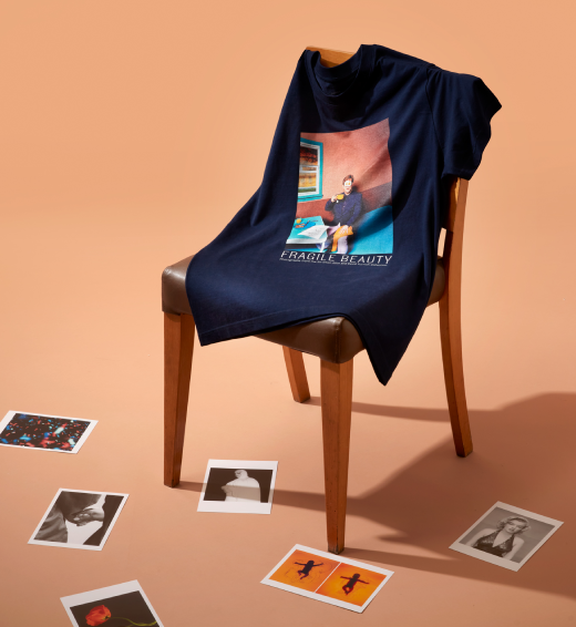 In an empty space, a chair sits strewn with a t-shirt featuring Elton John: Egg on His Face by David LaChapelle surrounded by scattered postcards of contemporary photography